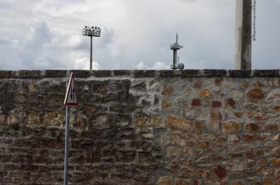 photograph “V.2014” par David Farreny — www.farreny.net — France, mur, wall, ville, city, pierre, stone, panneau, routier, sign, roadsign, flèches, arrows, triangle, poteaux, posts, poles, mâts, projecteurs, floodlights, paraboles, dish aerials, satellite dishes, dish antennas, métal, metal, nuages, clouds, échelle, ladder, Occitanie, Aveyron, Rouergue, Rodez