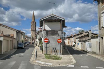 photograph “IX.2024 — Double bind” par David Farreny — www.farreny.net — France, Occitanie, Tarn-et-Garonne, Nègrepelisse, bourgade, town, rues, streets, parallèles, parallel, maisons, houses, bitume, blacktop, asphalte, asphalt, trottoir, pavement, sens interdit, one way, no entry, panneaux, signs, routiers, roadsigns, toit, roof, antenne, antenna, église, church, clocher, bell tower, ciel, sky, nuages, clouds, voitures, cars, femme, woman, tuiles, tiles, brique, brick, ciment, cement, herbe, grass, balcon, balcony, store, gouttière, drainpipe, métal, metal, portail, gate, poubelles, trash, cans, bins, ombre, shadow, paraboles, satellite, dishes