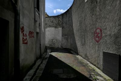 photograph “III.2022” par David Farreny — www.farreny.net — France, Occitanie, Ariège, Foix, rue, street, ruelle, alley, mur, wall, béton, concrete, tags, taches, stains, chien, dog, grille, railings, métal, metal, gouttière, drainpipe, portes, doors, fenêtre, window, bitume, blacktop, asphalte, asphalt, ciel, sky, ombre, shadow, peinture, paint, rouge, red, 1312, nuages, clouds