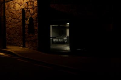 photograph “VIII.2023 — Another place you miss” par David Farreny — www.farreny.net — Andorre, Andorra, Canillo, nuit, night, soir, evening, sombre, dark, darkness, pénombre, noir, black, rue, street, trottoir, pavement, mur, wall, pierre, stone, garage, portail, gate, voiture, car, tarpaulin, tarp, sheet, plastique, plastic, béton, concrete, lumière, light, néon, neon, parcmètre, parking meter, intérieur, interior, bitume, blacktop, asphalte, asphalt