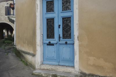 photograph “I.2025 — Faces of home, 252” par David Farreny — www.farreny.net — France, Occitanie, Gascogne, Gascony, Lomagne, Gers, Saint-Clar, rue, street, village, maison, house, porte, door, bois, wood, bleu, blue, poignée, handle, métal, metal, pierre, stone, sonnette, doorbell, mur, wall, beige, arcades, balcon, balcony, fleurs, flowers, pot, fenêtre, window, volets, shutters, marche, step, crépi, roughcast, gouttière, drainpipe, domicile, home