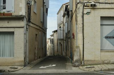 photograph “II.2025 — Painting” par David Farreny — www.farreny.net — France, Occitanie, Gascogne, Gascony, Lomagne, Gers, Lectoure, ville, city, bourgade, town, rue, street, carrefour, crossing, maisons, houses, pierre, stone, beige, vitrines, windows, fenêtres, verre, vitre, glass, angles, coins, corners, haut-parleur, loudspeaker, câbles, cables, fils, wires, bitume, blacktop, asphalte, asphalt, volets, shutters, bois, wood, panneaux, signs, métal, metal, rideau, curtain, flèche, arrow, lampadaires, lampes, lamps, enseigne, sign, peinture, painting, trottoir, pavement, plantes, plants, losange, diamond, perspective