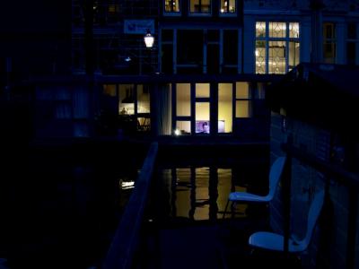 photograph “X.2017 — Faces of home, 95” par David Farreny — www.farreny.net — Pays-Bas, Netherlands, Nederland, Hollande, Holland, Amsterdam, canal, eau, water, soir, evening, chaises, chairs, péniche, barge, bateau, boat, maison, house, façade, facade, fenêtres, windows, téléviseur, télévision, TV, TV set, homme, man, écran, screen, lumières, lights, rideaux, curtains, noir, dark, pénombre, darkness, reflet, reflection, lampadaire, lamp