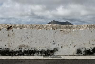 photograph “II.2018 — Lancerotte, 229” par David Farreny — www.farreny.net — Espagne, Spain, España, Canaries, Canary, Canarias, Lanzarote, Lancerotte, île, island, Teguise, volcan, volcano, montagne, mountain, blanc, white, mur, wall, paroi, rue, street, trottoir, pavement, bitume, blacktop, asphalte, asphalt, nuages, clouds