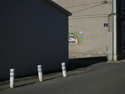 photograph “II.2025 — About the circus of living” par David Farreny — www.farreny.net — France, Occitanie, Tarn-et-Garonne, Septfonds, village, rue, street, maisons, houses, angles, coins, corners, carrefour, crossing, poteaux, plastique, plastic, posts, ombre, shadow, câbles, cables, fils, wires, gouttière, drainpipe, mur, wall, pierre, stone, enduit, plaque, plate, rue Jean Lacan, enseigne, sign, accueil de loisirs, leisure center, soleil, sun, bitume, blacktop, asphalte, asphalt, dépression, depression