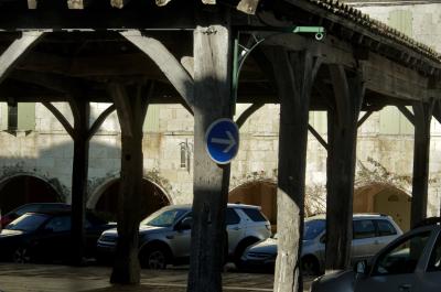 photograph “II.2025” par David Farreny — www.farreny.net — France, Occitanie, Gascogne, Gascony, Lomagne, Gers, Saint-Clar, village, place, square, halle, market hall, piliers, pillars, bois, wood, voitures, cars, métal, metal, arcades, archs, arcs, maisons, houses, pierre, stone, beige, panneau, sign, routier, roadsign, flèche, arrow, right, droite, fenêtres, windows, volets, shutters, tuiles, tiles