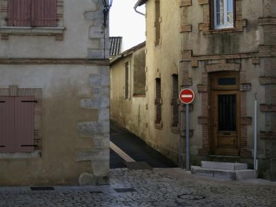 photograph “I.2025 — Exit route” par David Farreny — www.farreny.net — France, Occitanie, Tarn-et-Garonne, Caussade, ville, city, bourgade, town, rue, street, ruelle, alley, maisons, houses, lumière, light, bitume, blacktop, asphalte, asphalt, pavés, cobblestones, pierres, stones, fenêtres, windows, volets, shutters, bois, wood, marches, steps, lampadaire, lampe, lamp, gouttière, gutter, vitre, verre, glass, panneau, sign, routier, roadsign, sens interdit, one way, toit, roof, tuiles, tiles, brique, brick, étroit, narrow, plaque, plate, numéro, number, 26, échappée, escape, évasion