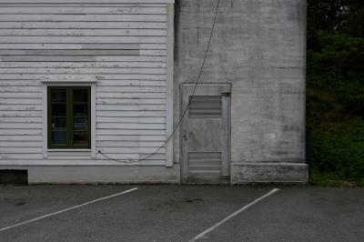 photograph “VI.2019 — Run away to the North, 180” par David Farreny — www.farreny.net — Norvège, Norway, Norge, Hordaland, Austevoll, Bekkjarvik, parking, car park, bâtiment, building, maison, house, béton, concrete, bois, wood, planches, planks, bitume, blacktop, asphalte, asphalt, fenêtre, window, fil, wire, câble, cable, porte, door, gris, grey, gray