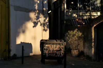 photograph “VII.2022” par David Farreny — www.farreny.net — France, Provence-Alpes-Côte d'Azur, PACA, Bouches-du-Rhône, Aix-en-Provence, été, summer, soir, evening, rue, street, poubelle, conteneur, trash, can, container, graffiti, Jules Pneus, feuillage, foliage, ombre, tree, mur, wall, plante, plant, pot, poteaux, posts, poles, métal, metal, plastique, plastic, escalier, stairs, staircase, marches, steps, verre, vitre, glass