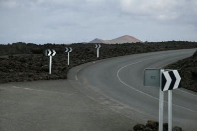 photograph “II.2018 — Lancerotte, 82” par David Farreny — www.farreny.net — Espagne, Spain, España, Canaries, Canary, Canarias, Lanzarote, Lancerotte, île, island, volcan, volcano, montagne, mountain, bitume, blacktop, asphalte, asphalt, noir, black, route, road, rochers, rocks, lave, lava, nuages, clouds, virage, turn, panneaux, signs, métal, metal, flèches, arrows, droite, right