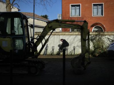 photograph “I.2025” par David Farreny — www.farreny.net — France, Occitanie, Tarn-et-Garonne, Caussade, ville, city, bourgade, town, tractopelle, backhoe loader, véhicule, travaux, works, voirie, road, rue, street, bâtiment, immeuble, building, trésor, treasury, fisc, tax, services, centre des Finances publiques, Public Finance Center, vol légal, legal theft, logo, F, fenêtres, windows, mur, wall, brique, brick, arbre, tree, toit, roof, jeune femme, young woman, assise, sitting, poteau, pole, métal, metal, voiture, car, verre, vitre, glass, téléphone, phone, smartphone, penchée, leaning over, marque, brand, Yanmar, pelle, shovel, cabine, cabin, branches, plantes, plants, lévitation, levitation