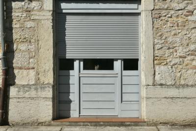 photograph “IX.2024 — Watcher” par David Farreny — www.farreny.net — France, Occitanie, Tarn-et-Garonne, Septfonds, maison, house, mur, wall, paroi, pierre, stone, gouttière, drainpipe, métal, metal, porte, door, store, bois, wood, ombre, shadow, chat, cat, animal