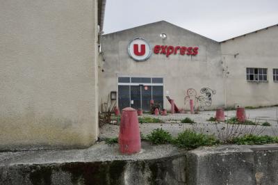 photograph “I.2025” par David Farreny — www.farreny.net — France, Occitanie, Gascogne, Gascony, Gers, Mauvezin, supermarché, supermarket, fermé, closed down, entrée, entrance, logo, enseigne, sign, U Express, béton, concrete, ciment, cement, plots, blocks, rouge, red, tag, graffiti, crépi, roughcast, fenêtres, windows, porte, door, vitre, verre, glass, grille, grid, abandonné, abandoned, fleurs, flowers, lampes, lamps, gouttière, gutter, métal, metal