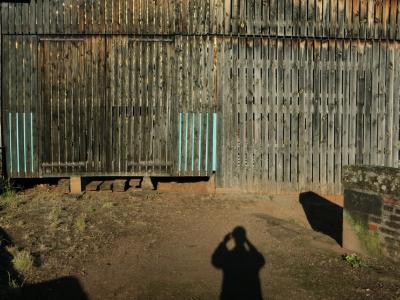 photograph “VI.2012 — Hatched self” par David Farreny — www.farreny.net — France, soir, evening, grange, barn, bois, wood, planches, planks, paroi, mur, wall, ombre, shadow, autoportrait, selfportrait, hachures, hatching, parapet, pierre, stone, brique, brick, terre, earth, sol, soil, silhouette, figure, Occitanie, Rouergue, Aveyron, Marcillac-Vallon