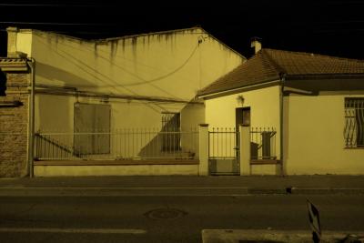 photograph “X.2022 — Faces of home, 239” par David Farreny — www.farreny.net — France, Occitanie, Haute-Garonne, Toulouse, nuit, night, maison, house, toit, roof, clôture, grille, fence, rue, street, panneau, sign, flèche, arrow, fenêtres, windows, porte, door, câbles, cables, fils, wires, ombres, shadows, trottoir, pavement, bitume, blacktop, asphalte, asphalt, ciment, cement, brique, brick, gouttière, drainpipe, jaune, yellow, lumière, light