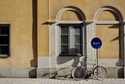 photograph “VIII.2019 — Run away to the North, 272” par David Farreny — www.farreny.net — Suède, Sverige, Sweden, Uppland, Södermanland, Stockholm, ville, city, capitale, capital, Södermalm, mur, wall, fenêtres, métal, metal, pierre, stone, verre, vitre, glass, bicyclette, bicycle, vélo, bike, pavés, cobblestones, arches, arcs, archs, panneau, sign, routier, roadsign, flèche, arrow, gauche, left, ombres, shadows