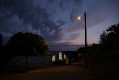 photograph “IX.2019 — Municipal achievements, 8. Sports hall” par David Farreny — www.farreny.net — France, gymnase, gymnasium, gym, salle omnisports, sport hall, soir, evening, automne, autumn, fall, lampadaire, réverbère, lamp, rue, street, arbres, trees, lumière, light, ciel, sky, nuages, clouds, tôle ondulée, corrugated iron, métal, metal, laid, ugly, bâtiment, building, village, campagne, countryside, Occitanie, Gascogne, Gers, Jegun