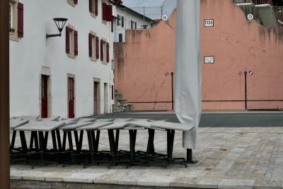 photograph “XII.2020” par David Farreny — www.farreny.net — France, Aquitaine, Pyrénées-Atlantiques, Bidart, Labourd, Pays basque, place, square, fronton, court, pelote basque, Basque pelota, maisons, houses, pierre, stone, dalles, paving, flagstones, tables, parasol, sunshade, fenêtres, windows, volets, shutters, bois, wood, drapeaux, flags, français, French, Basque, ikurrina, dates, années, years, year, 1925, 1869, 1912, croix basque, Basque cross, lauburu, svastika, lampe, lamp, grillage, fence, bitume, blacktop, asphalte, asphalt, métal, metal, toile, canvas, bâche, sheet, Basque country