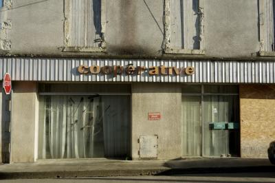 photograph “I.2025” par David Farreny — www.farreny.net — France, Occitanie, Gascogne, Gascony, Lomagne, Gers, Fleurance, bâtiment, building, rue, street, enseigne, coopérative, cooperative, vitrine, window, verre, vitre, glass, rideaux, curtains, fermé, closed down, fenêtres, windows, volets, shutters, bois, wood, vieux, old, pierre, stone, trottoir, pavement, panneau, routier, roadsign, stop, crépi, roughcast, stationnement interdit, no parking, ombre, shadow, porte, door