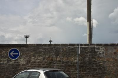 photograph “V.2014 — Sunday” par David Farreny — www.farreny.net — France, Occitanie, Aveyron, Rouergue, Rodez, stade, stadium, mur, wall, pierre, stone, panneau, sign, routier, roadsign, flèche, arrow, left, gauche, poteaux, posts, poles, projecteurs, floodlights, voiture, car, métal, metal, ciel, sky, nuages, clouds, échelle, ladder, reflet, reflection, vitre, verre, glass, dépression, depression