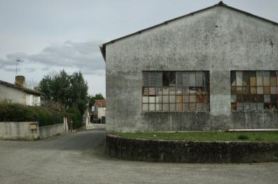 photograph “II.2025 — Whatever” par David Farreny — www.farreny.net — France, Occitanie, Gascogne, Gascony, Lomagne, Gers, Saint-Clar, village, entrepôt, warehouse, hangar, shed, ciment, cement, baies, fenêtres, windows, verre, vitre, glass, accumulation, objets, things, toit, roof, gouttière, gutter, rue, street, herbe, grass, maisons, houses, bitume, blacktop, asphalte, asphalt, haie, badge, arbres, trees, boîte à lettres, mailbox, métal, metal, antenne, antenna, cheminée, chimney, volets, shutters, nuages, clouds